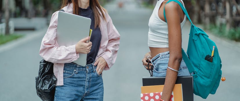 Crop anonymous multiethnic female students with backpacks and device with textbooks standing on footpath in park
